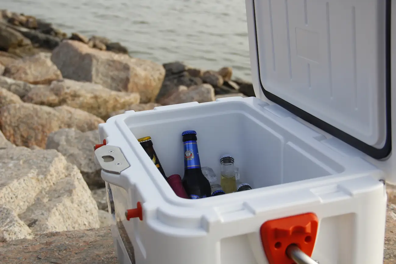 Hielera blanca abierta con bebidas frías, junto a unas rocas y el agua